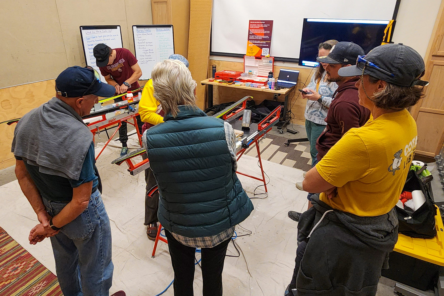 Participants gather around a workbench during the CMU Cross Country Ski Team’s free waxing clinic at REI Grand Junction, watching as a student-athlete demonstrates ski waxing techniques using Swix tools and equipment.
