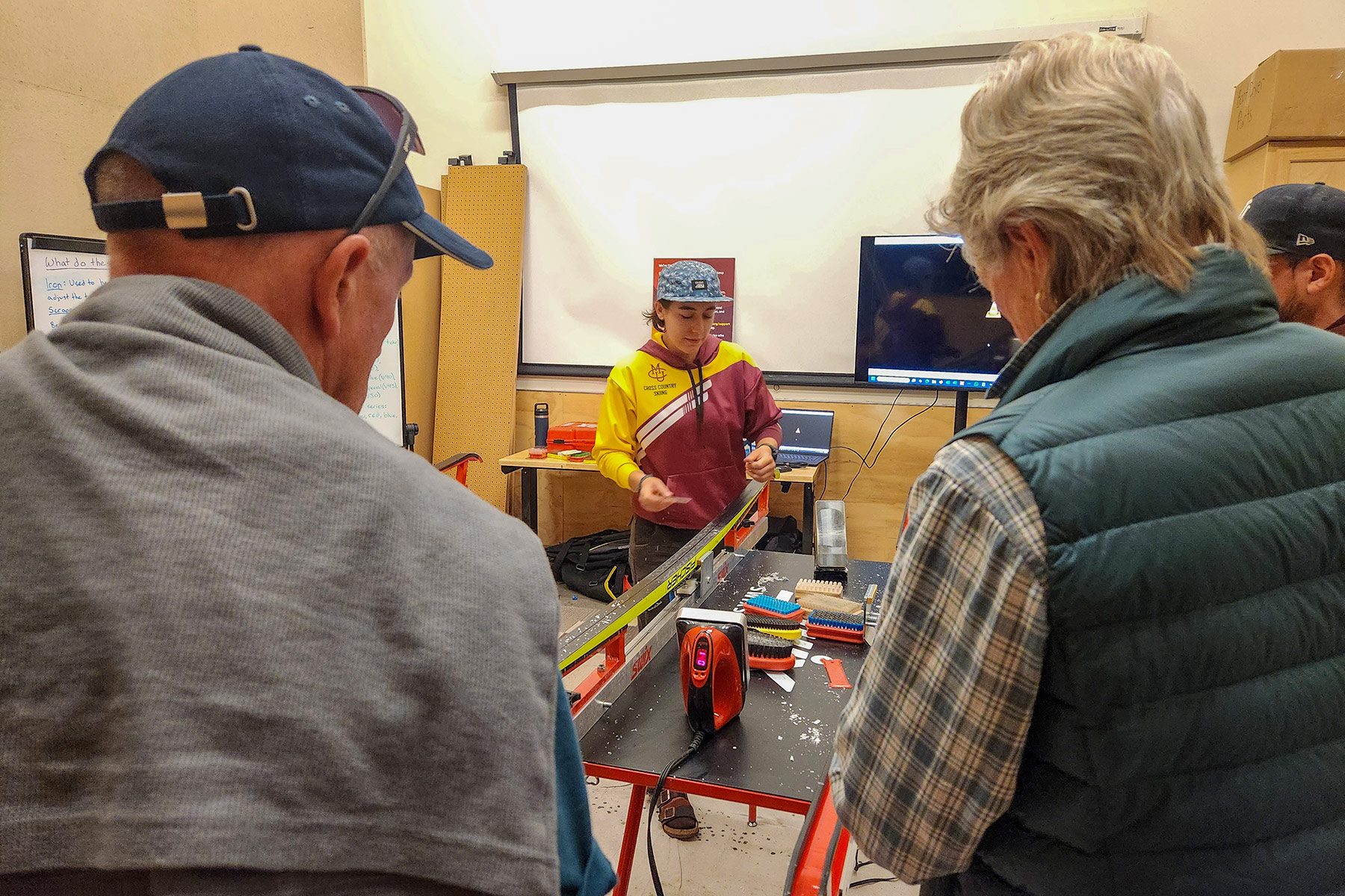 A CMU Cross Country Ski Team athlete demonstrates how to wax cross country skis during a free community clinic at REI Grand Junction, with participants closely observing the process.