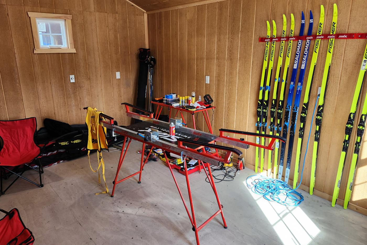 Cross-country ski waxing room with Swix ski benches, waxing tools on a work table, Fischer and Salomon skis stored on a wall rack, and folding chairs along the side.