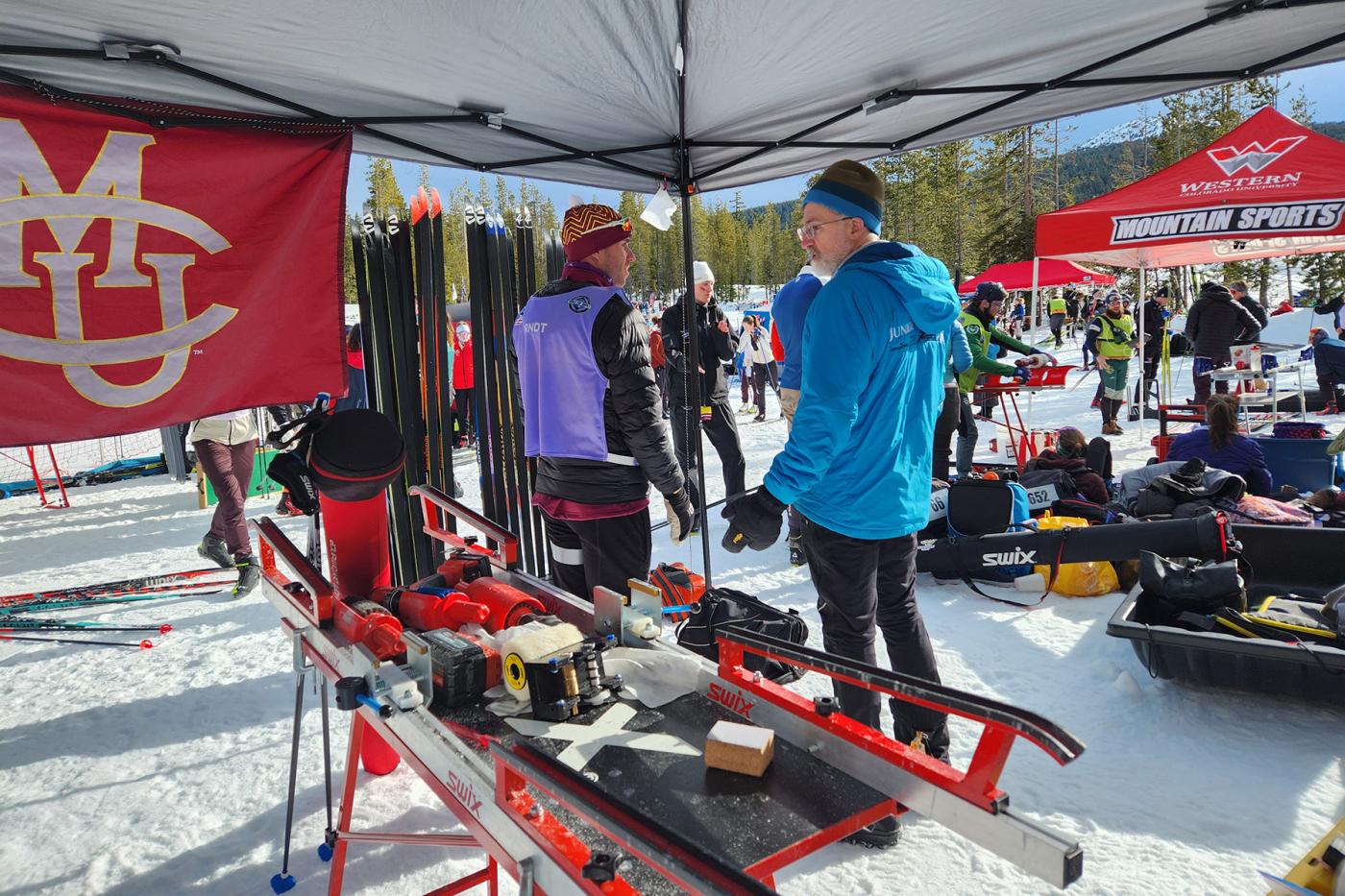 Ski waxing station under a team tent at a cross-country ski race, with people preparing skis and equipment in the snow.