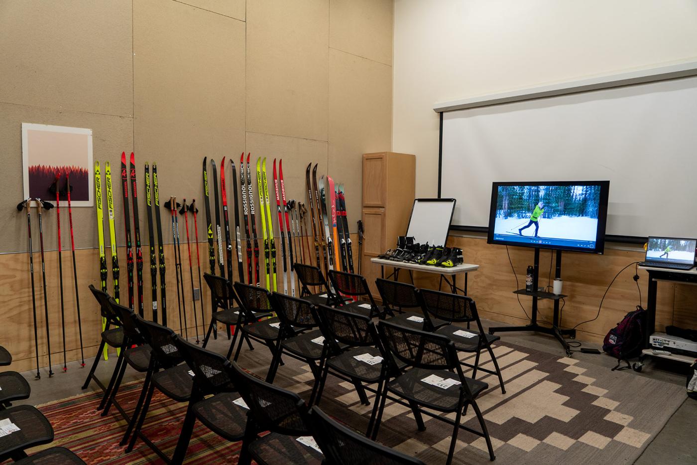 Classroom setup at REI for a ski gear clinic with rows of chairs facing a screen, cross-country skis and poles lined up along the wall, ski boots on a table, and a video of skiing playing on a monitor.