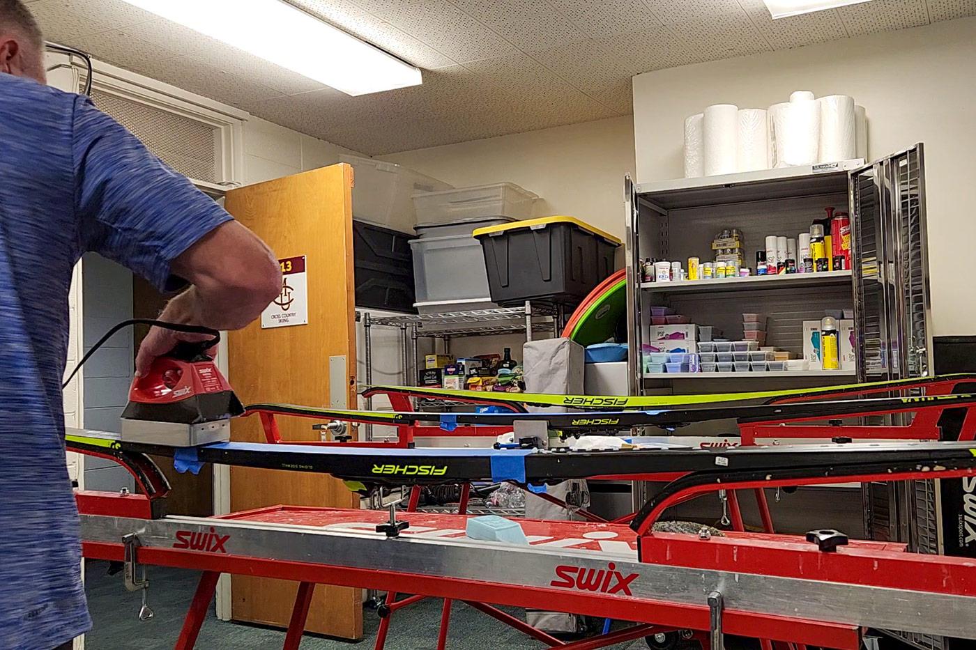 Person waxing cross-country skis with a Swix waxing iron in a ski prep room, with skis secured in Swix benches and shelves stocked with ski waxes and tools in the background.