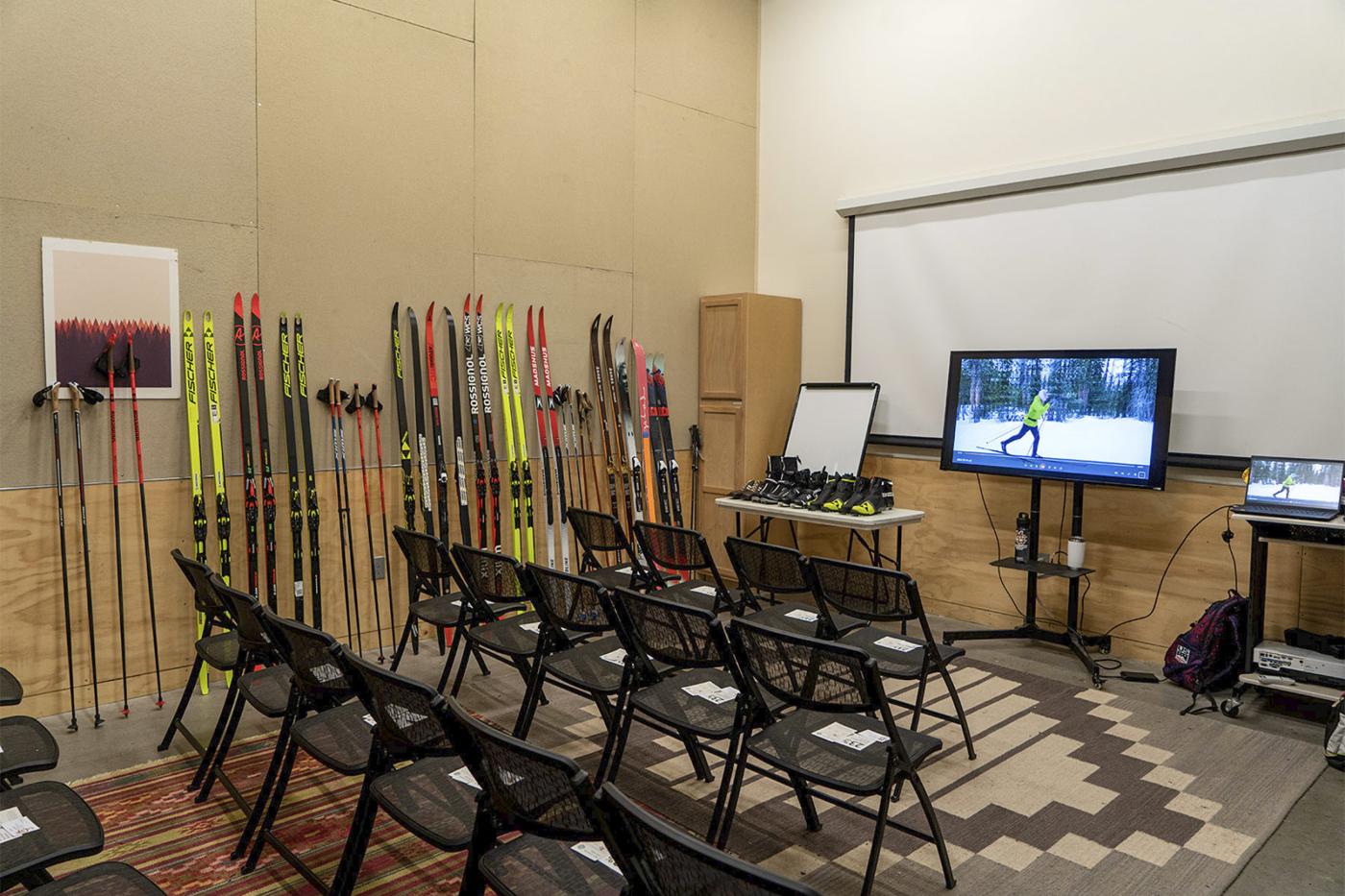 Classroom setup at REI for a ski gear clinic with rows of chairs facing a screen, cross-country skis and poles lined up along the wall, ski boots on a table, and a video of skiing playing on a monitor.