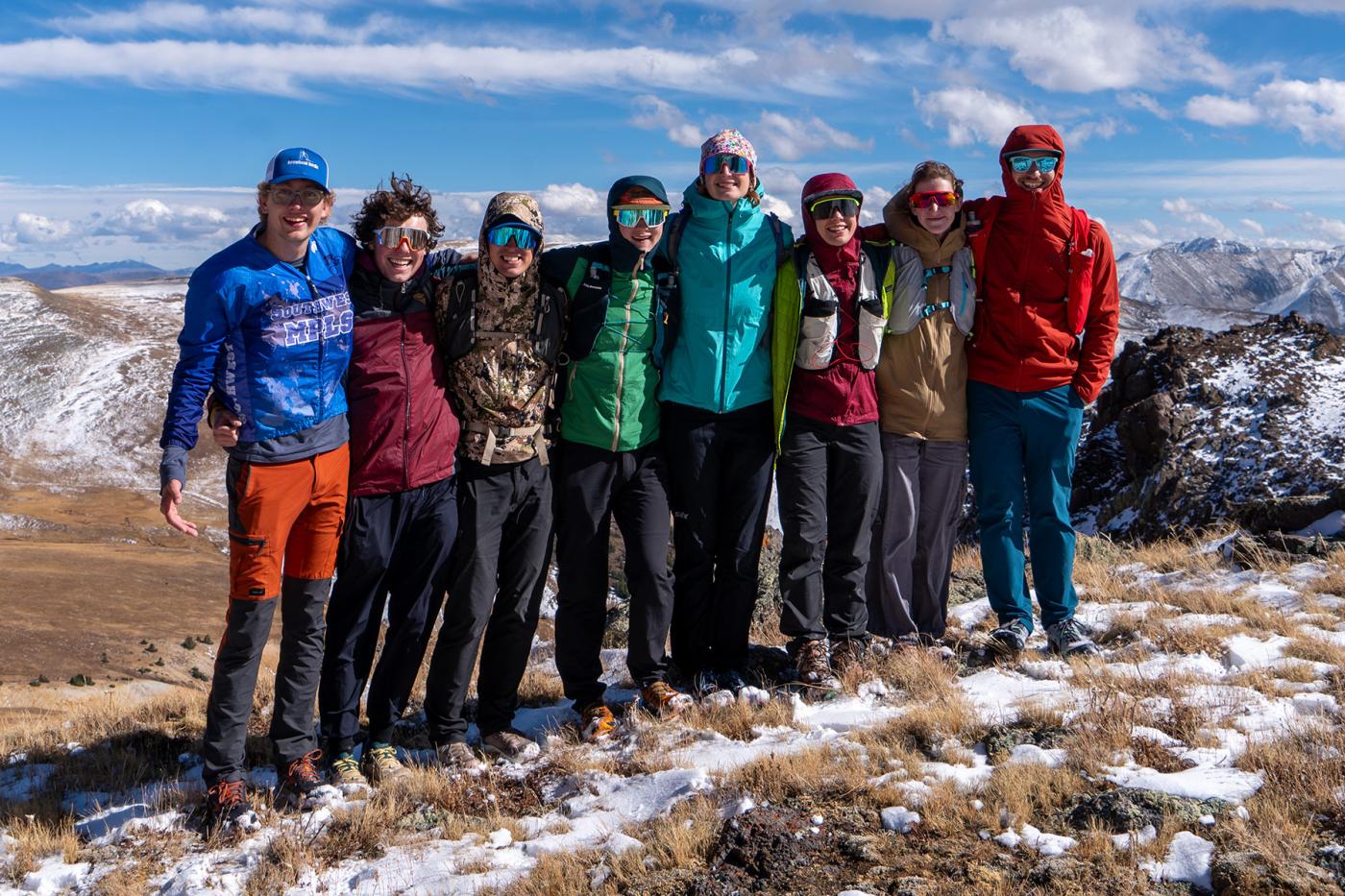 Colorado Mesa University Cross Country Ski Team athletes standing together on a high alpine ridge with snow-dusted ground and mountain peaks in the background, smiling and wearing colorful jackets and sunglasses on a bright, windy day.